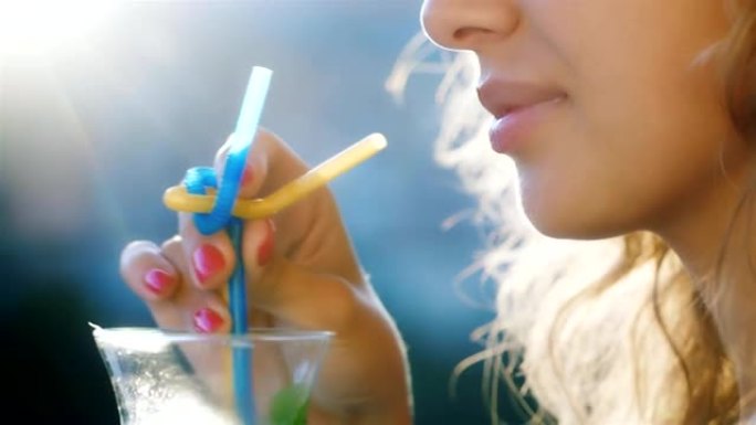 年轻的女人在杯子里搅拌饮料。在图片中，可见阳光照耀的嘴唇和头发。概念-天堂般的假期