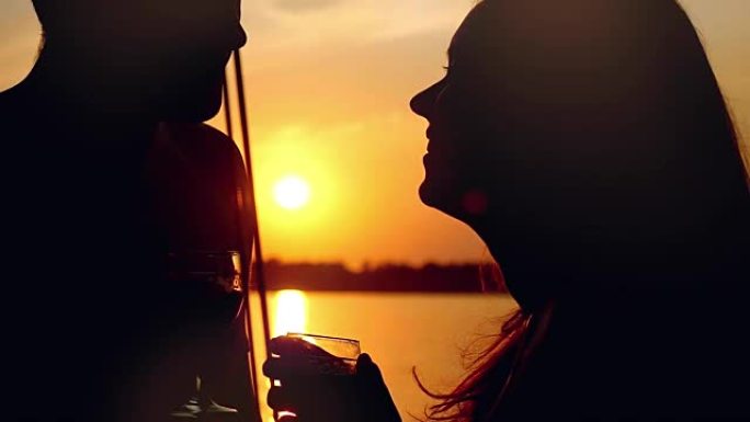 在太阳背景下缓慢运动的日落时，在帆船上恋爱的年轻夫妇带着一杯葡萄酒