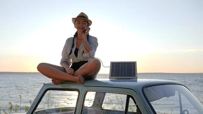 生态旅游，年轻女子使用太阳能电池在背景蓝天上移动，坐在阳光下的老式汽车上