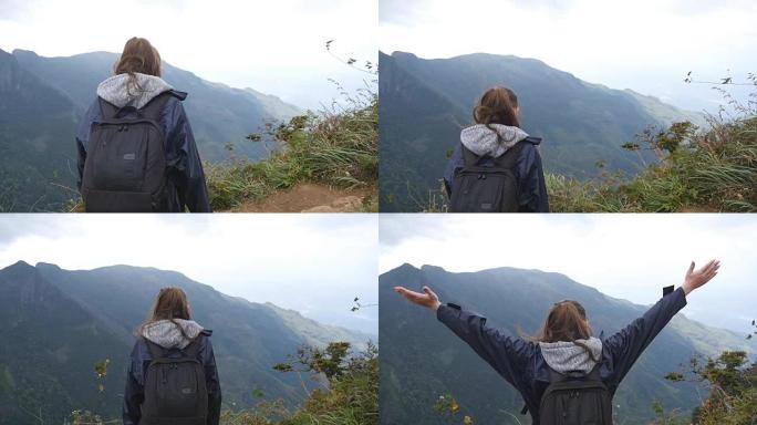 年轻的女徒步旅行者，背包爬到山顶，举起双手。穿着雨衣的女游客站在美丽的峡谷边缘，胜利地伸出双臂。慢动
