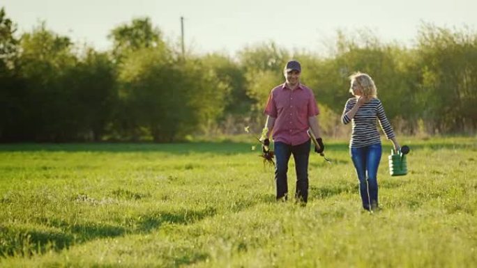 一对年轻的农民夫妇沿着一片绿地附近的乡间小路行走。携带树苗和喷壶。种树，在农村工作