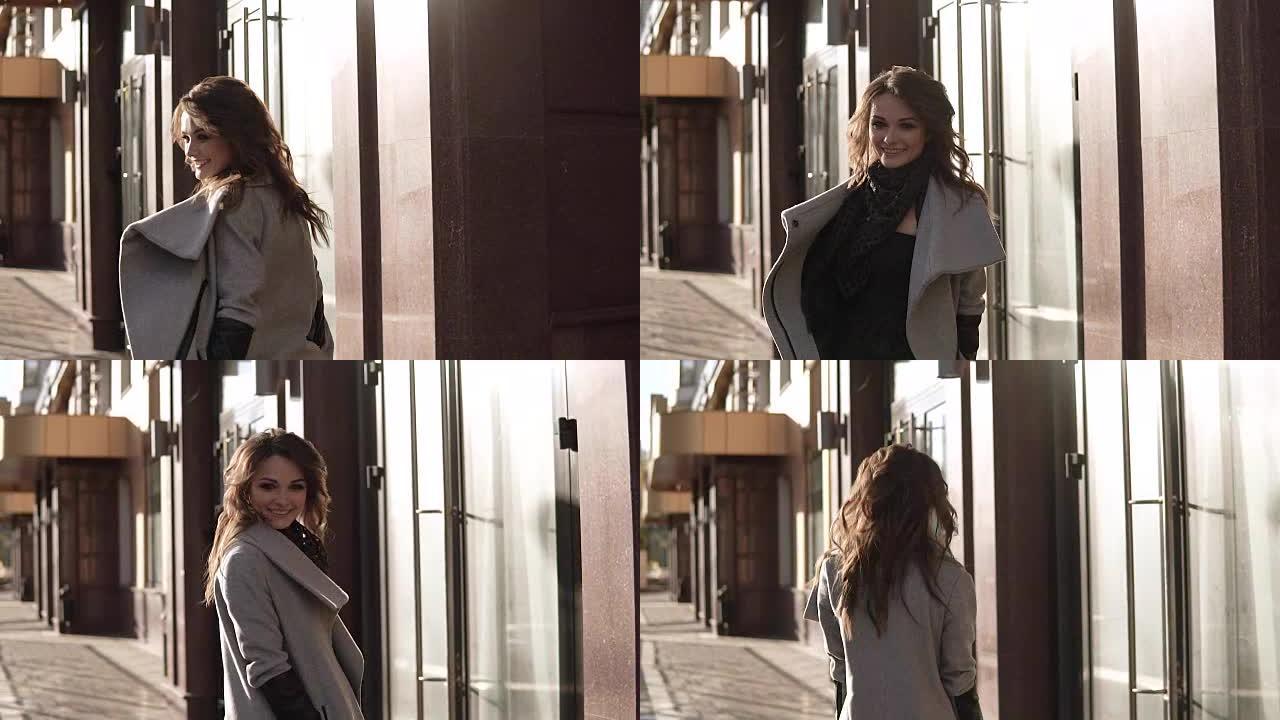 迷人的年轻女子穿着外套走过秋天的城市，微笑着。现代建筑背景下的可爱女孩。慢动作