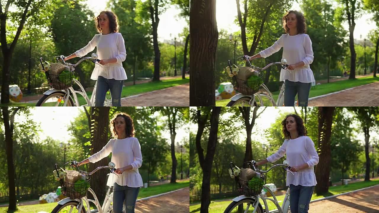 穿着白色衬衫和蓝色牛仔裤的迷人女人走在城市公园的小巷上，手里拿着城市自行车的车把，篮子里放着鲜花。透