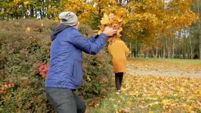 父亲和女儿在秋季公园玩耍。然而，他们微笑着笑。爸爸把树叶扔在一个少年的头上