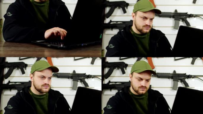 倾斜射击:一个年轻人正在用笔记本电脑工作，背景是一个有步枪和其他武器的支架