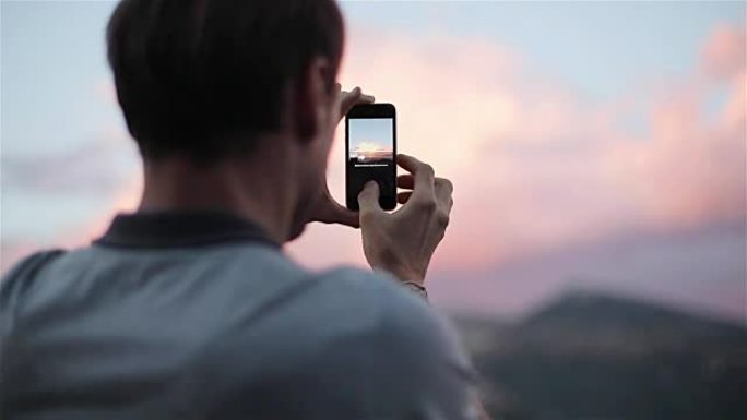 Man traveller使全景照片保持和转动手机站在日落山的顶部慢动作从背面和侧面关闭。科技旅行快