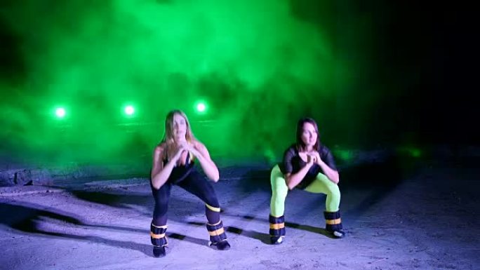 两名运动妇女，在晚上，在轻烟，雾中，在五彩缤纷的探照灯下，在一个废弃的旧机库中，在腿上进行各种健身运