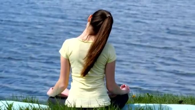 年轻女子做着做着头来回弯曲的动作。女孩坐在草地上，在河边冥想，在莲花姿势时做瑜伽练习