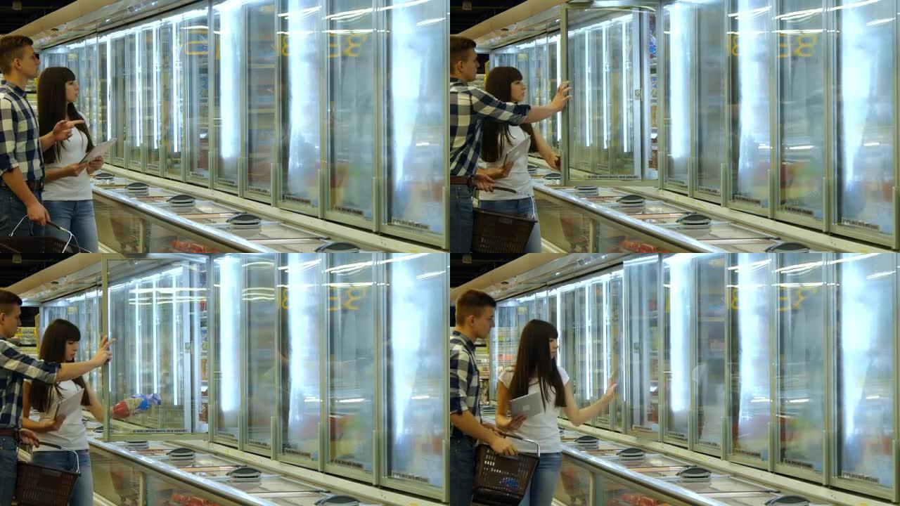 年轻夫妇站在杂货店的冰柜旁边，挑选装有冷冻食品的包装。男子从冰箱里拿走产品。女人使用平板电脑检查购物