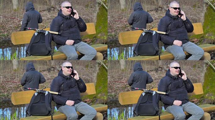 背着背包和钓鱼竿的男人坐在长凳上，在电话上聊天