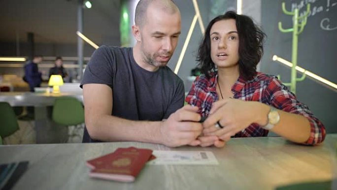 年轻夫妇坐在桌子旁，用手指点击智能手机屏幕。男人和女人在小工具上查看他们的照片，同时笑着并积极评论他