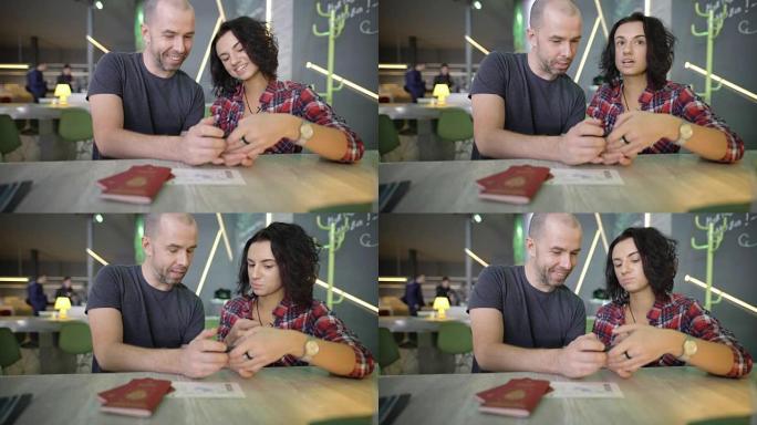 年轻夫妇坐在桌子旁，用手指点击智能手机屏幕。男人和女人在小工具上查看他们的照片，同时笑着并积极评论他