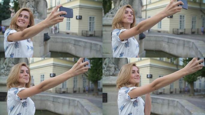 年轻女孩坐在城市喷泉上的智能手机自拍。快乐的年轻女子微笑着用苹果智能手机相机摆姿势自拍。旅游、ins