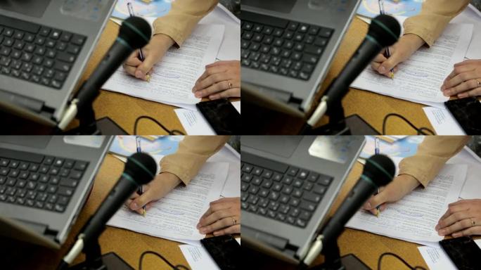 笔记本电脑和麦克风背景下的女性手在纸上做笔记和更正