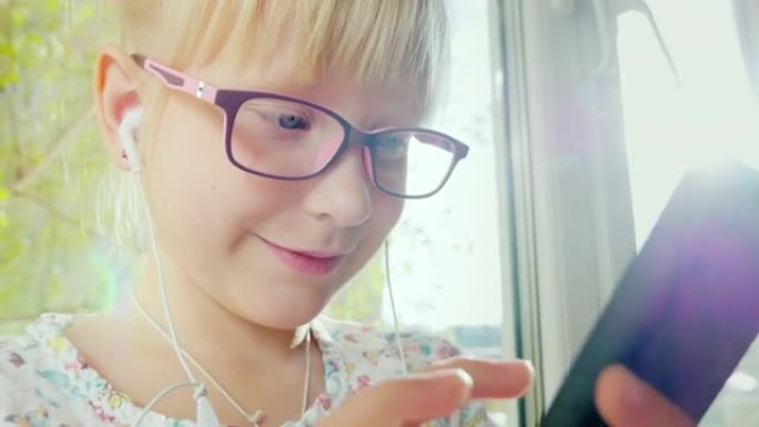 有趣的女孩微笑着使用电话。她戴着玫瑰色的眼镜和白色的耳机。一个带着设备的快乐孩子的肖像