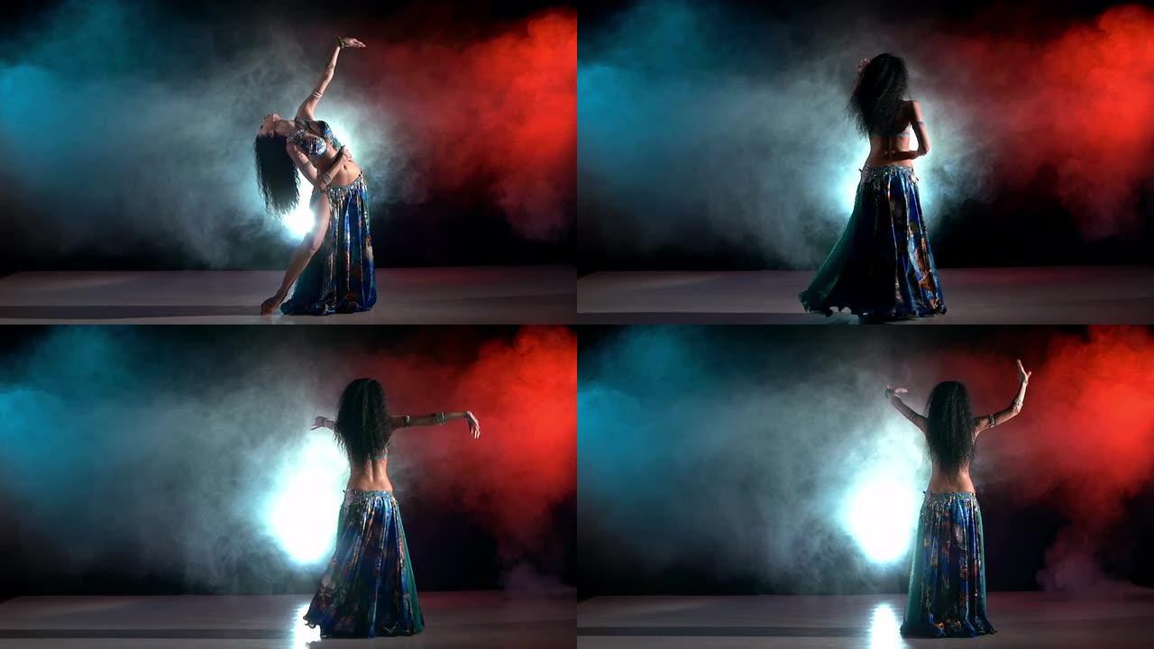 穿着蓝色异国情调服装的美丽年轻肚皮舞者继续跳舞，现代，黑色，红色，烟雾，慢动作