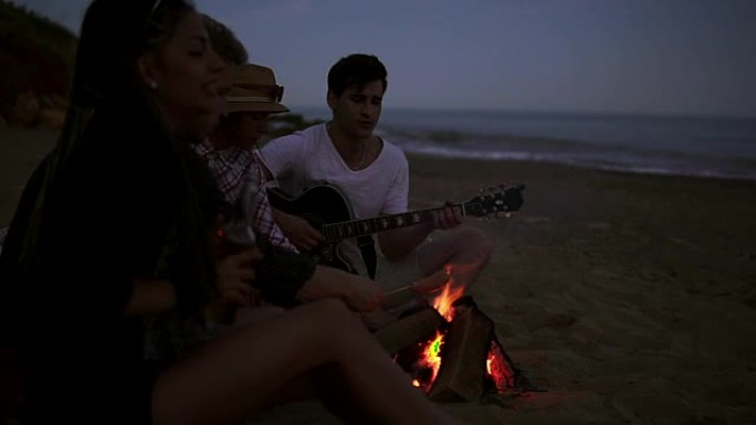 晚上，一群年轻开朗的人坐在海边的火炉旁，烤香肠和弹吉他。慢动作镜头