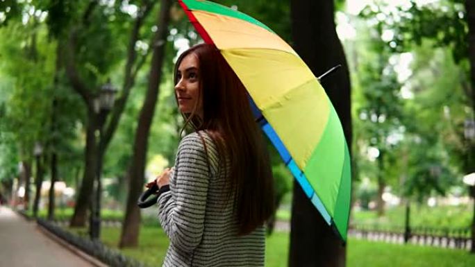 在城市公园的雨天，一个迷人的女人在旋转五颜六色的雨伞的后景