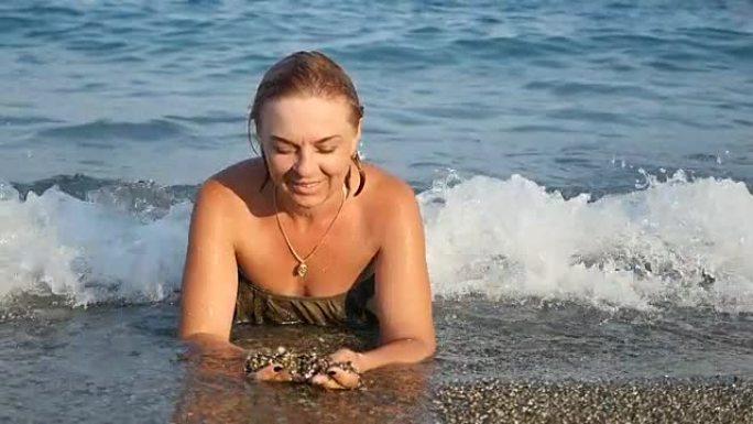 年轻女子躺在整理海水的海滩上，在slo-mo中选择贝壳