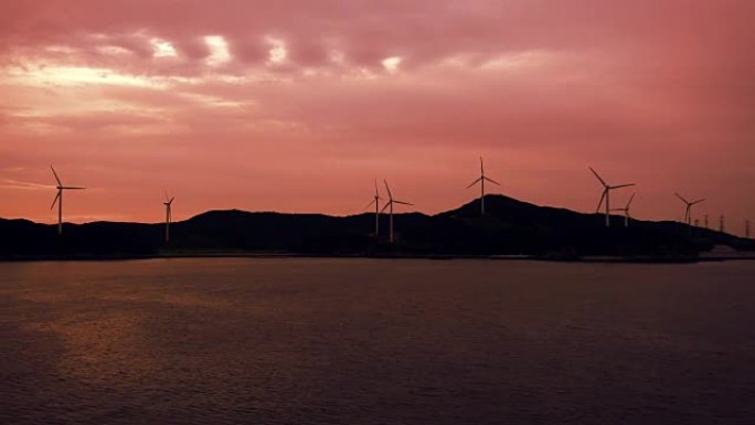 在暴风雨的海洋海岸线上，发电站和风力发电机的轮廓在日出或日落时带有辐射的火焰云。