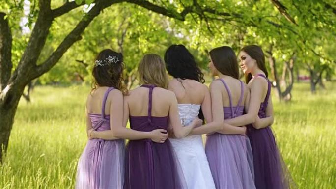从情感新娘和伴娘的背后看，他们在说话和微笑。穿着紫色连衣裙的高加索女孩在户外摆姿势。