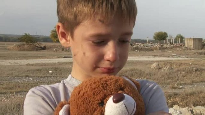 肮脏的小孤儿男孩特写哭泣抚摸毛绒玩具擦拭被毁房屋废墟背景上的眼泪