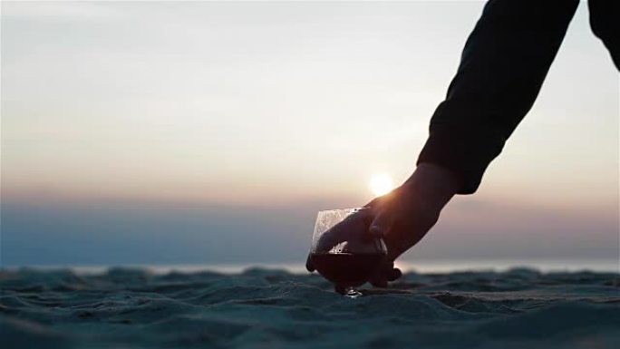 特写两杯白兰地站在沙滩上令人难以置信的海滩日落背景视图。男性的手勇敢地拿走眼镜，然后放回沙面