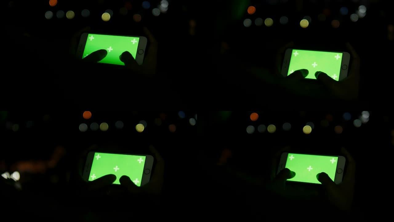 在具有散景背景的智能手机的色键绿色屏幕上玩游戏时，女人的手的特写-休闲活动的夜生活