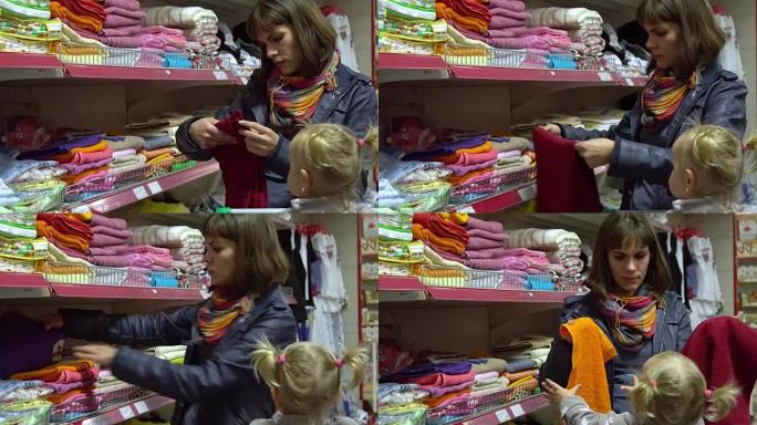 一个带着孩子在超市里的年轻女子选择商品。妈妈在手推车上开车送一个小女孩。妈妈和女儿选择毛巾。