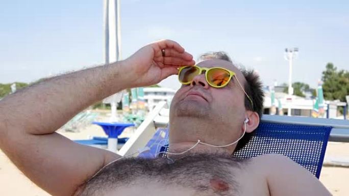 暑假、科技和人的概念 -- 戴着太阳镜的快乐年轻人，戴着智能手机和耳机听音乐，在沙滩巾上晒日光浴