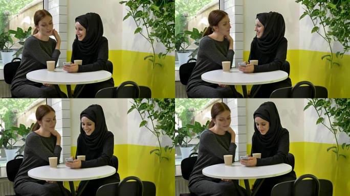 两个年轻漂亮的穆斯林妇女坐在咖啡馆里，其中一个戴着头巾，看着电话，笑着