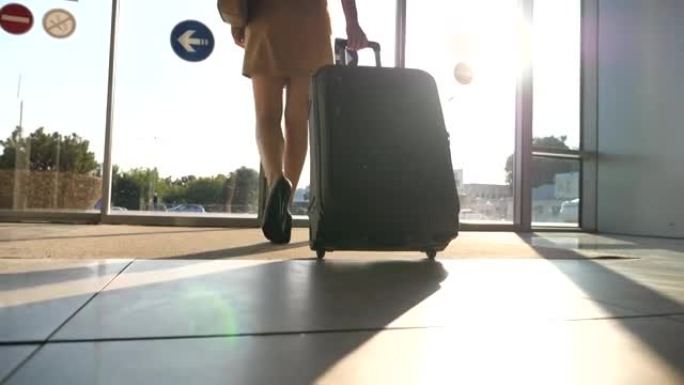 商务小姐带着行李从机场走。穿着高跟鞋的年轻女子穿过玻璃门走到街上，用轮子滚动她的手提箱。背景下的太阳