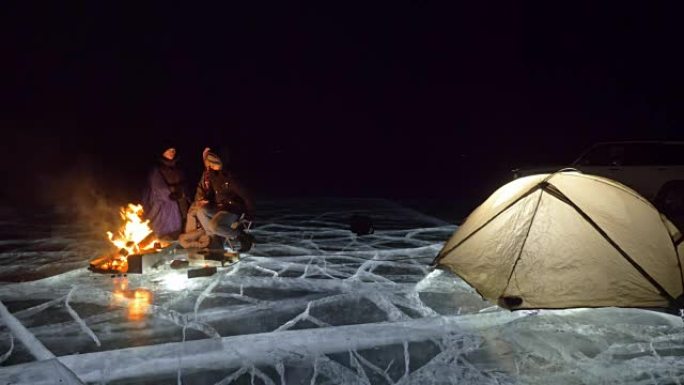 三个旅行者晚上在冰上着火。冰上的露营地。帐篷站在火旁边。贝加尔湖。附近有车。人们在篝火旁变暖，穿着睡