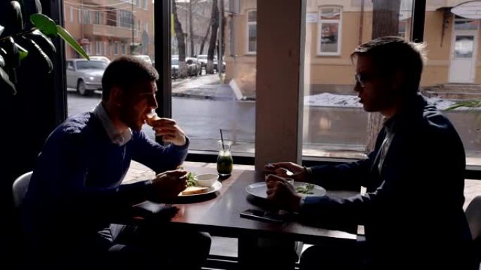 年轻的商人正在吃东西，坐在咖啡馆的餐桌旁，两个男人在聊天，就项目进行对话，在现代室内靠窗户享受美味的