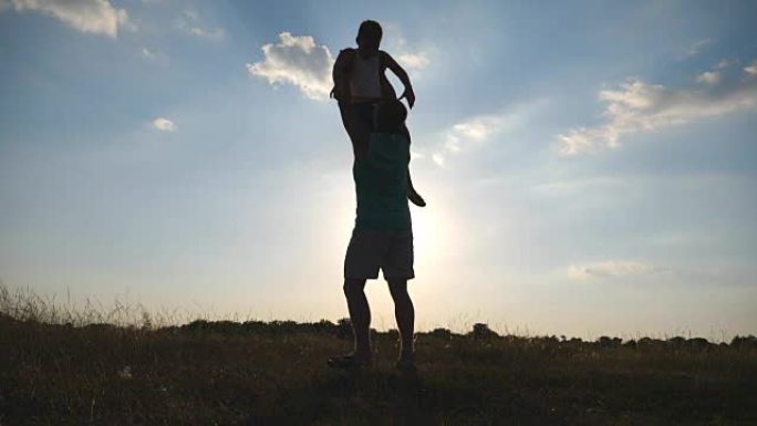 快乐爸爸的剪影把他的小男孩扔在草地上。父亲和儿子在日落时在球场上玩耍。爸爸和孩子在一起玩得开心。慢动