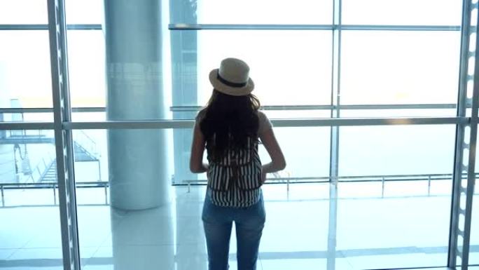 戴着帽子的年轻女子带着背包来到机场航站楼的窗口，看着跑道。女孩在等她的飞机。旅游和旅游概念。后视图特