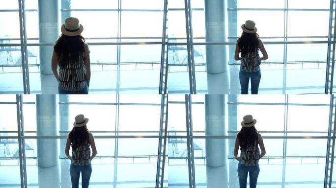 戴着帽子的年轻女子带着背包来到机场航站楼的窗口，看着跑道。女孩在等她的飞机。旅游和旅游概念。后视图特