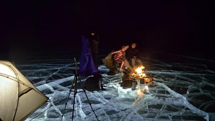 四个旅行者晚上在冰上着火。冰上的露营地。帐篷站在火旁边。人们在篝火旁变暖。摄影师在三脚架上拍摄。