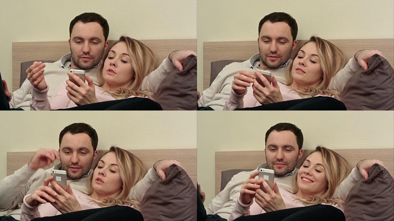 美丽的夫妇坐在床上，用智能手机触摸屏幕看旅行中的照片