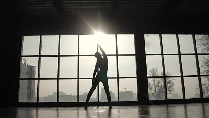 一个大窗户背景下运动员的轮廓。运动女孩在训练前做热身。练习瑜伽的年轻女子