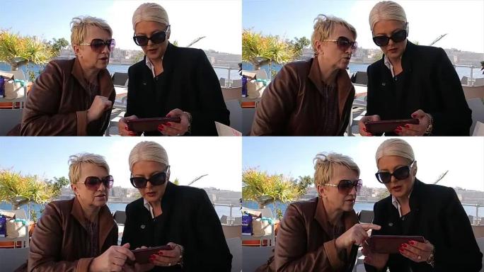 两个现代的成熟的成年妇女在海边的咖啡店聊天和使用手机在网上购物