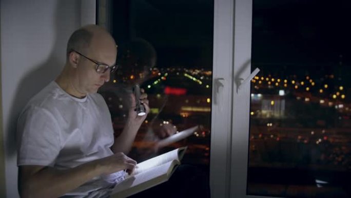 成年男子坐在窗台上，在黑暗中用手电筒浏览书页