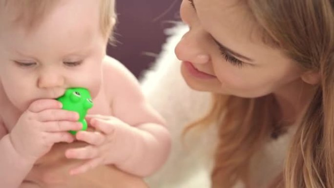 小婴儿坐在妈妈的怀里。婴儿在尿布吃绿色玩具
