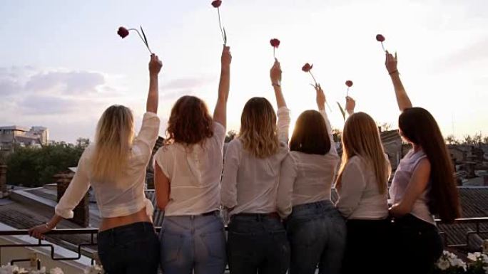 六名诱人的年轻女性从她们的背上连续站在露台上。穿着便服，白衬衫和牛仔裤。用一朵花举起他们的手。前视图