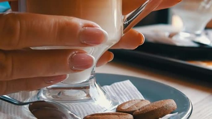 玻璃烧杯配咖啡，从深棕色分级为乳白色。咖啡馆里有很大泡沫的漂亮拿铁。一个女人用热拿铁触摸玻璃杯