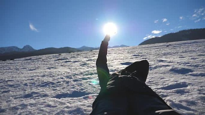 在阳光明媚的日子里，无法辨认的人躺在雪地上，用手指看着灿烂的阳光。年轻人在蓝天背景上触摸阳光的手。享