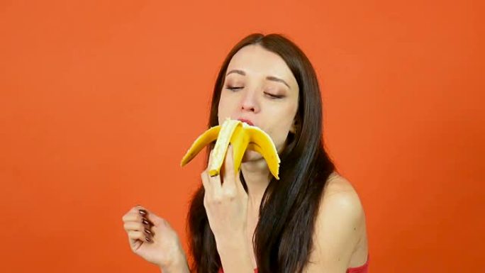 年轻健康的女人，皮肤完美，在明亮的橙色背景下吃香蕉。热带水果。素食概念