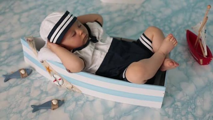 可爱的蹒跚学步的男婴睡在带鱼、海星和双筒望远镜的木船上，打扮成水手，在室内拍摄。