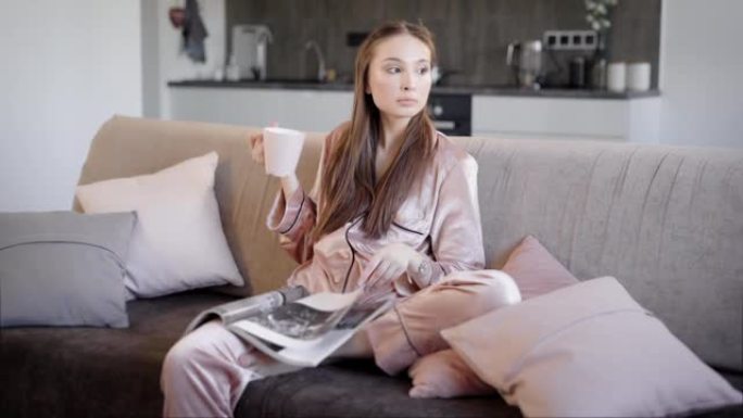 穿着自制睡衣的年轻女子坐在沙发上看时尚杂志，喝热茶