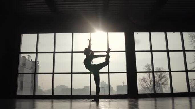 一个大窗户背景下运动员的轮廓。运动女孩在训练前做热身。练习瑜伽的年轻女子